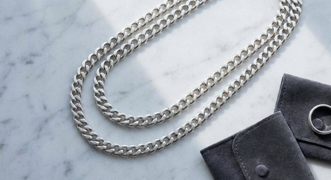 Two polished sterling silver curb chain necklaces of different widths laid side by side on a white marble surface, photographed from above.